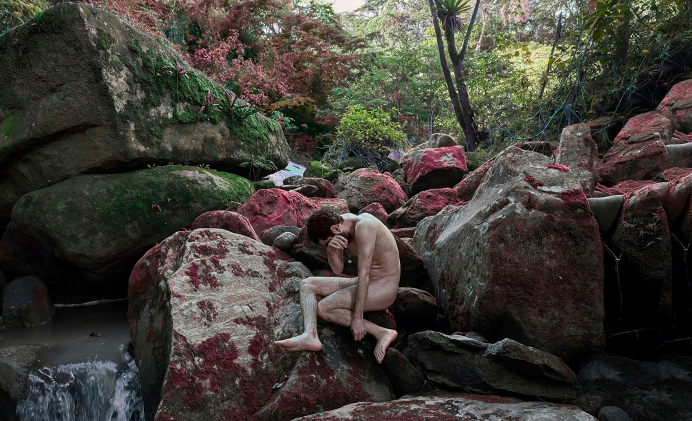 Artistic photograph in a forest, with a long-haired woman dressed in a long white dress, sitting on a giant rock, covering her face which is against her knees. On the other side there is a waterfall.